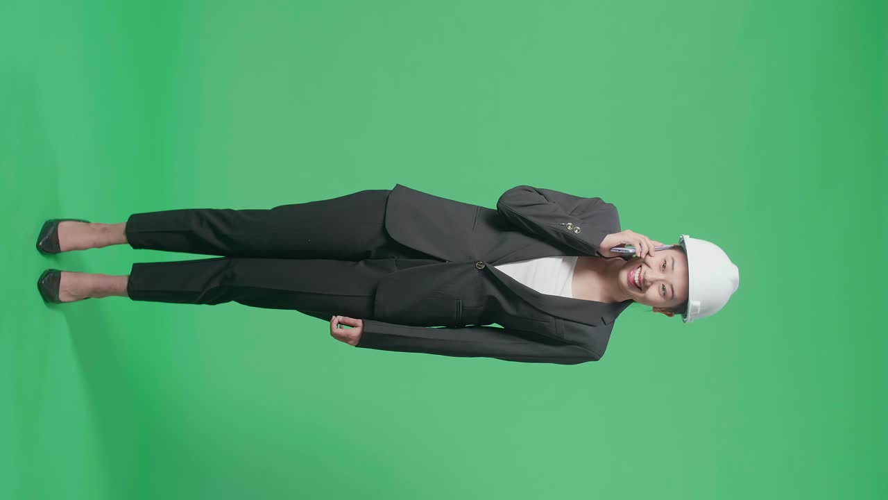 Full Body Of Smiling Asian Female Engineer With Safety Helmet Talking On Smartphone In The Green Screen Studio