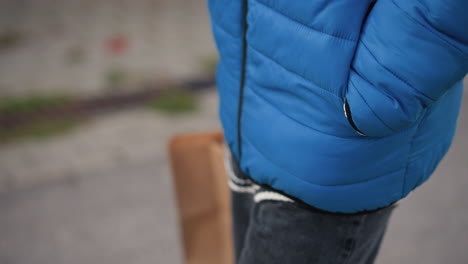 close-up of an individual's arm and side, wearing a bright blue puffer jacket and holding a shopping bag, with blurred urban background featuring greenery by the wayside