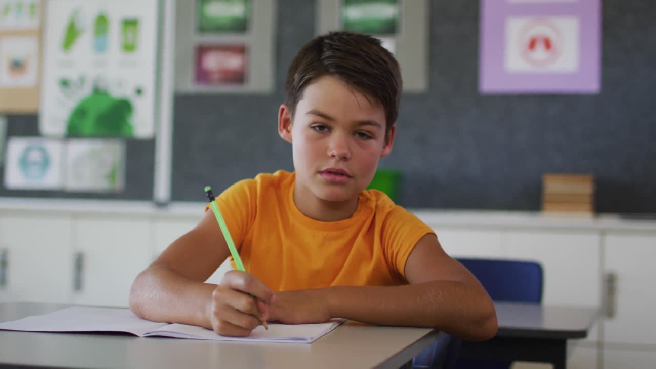 Retrato de un feliz escolar mestizo sentado en el aula, tomando apuntes y mirando a la cámara.