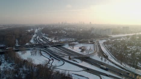 Imágenes-Aéreas-Establecidas-Que-Avanzan-En-Una-Carretera-De-Intersección-De-Tráfico-Cubierta-De-Nieve-Con-El-Horizonte-Del-Centro-De-La-Ciudad-De-Varsovia-Al-Atardecer-En-Invierno