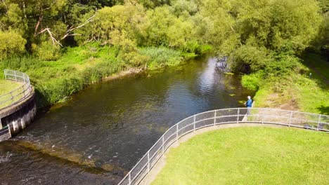 Aerial-ascending-shot-of-a-fisherman-standing-on-a-bank-of-the-small-water-dam-on-the-Little-Ouse-river-near-Thetford-in-the-UK