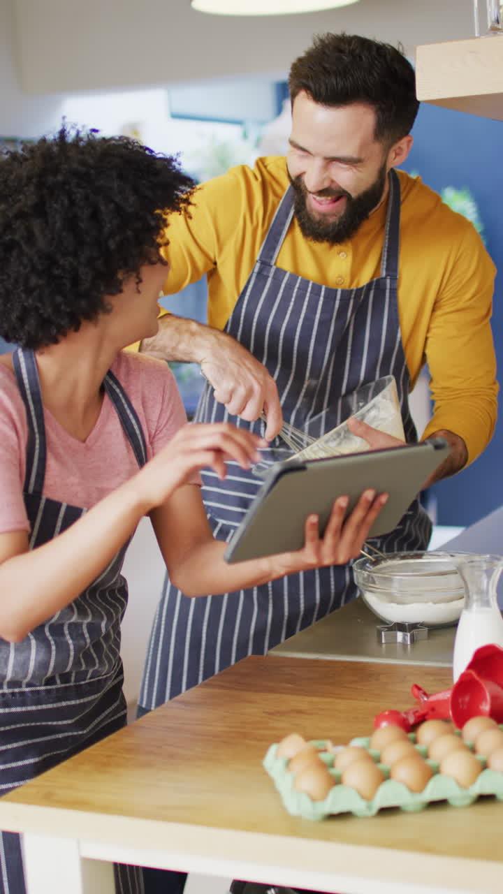 Vertical video of happy diverse couple baking in kitchen using tablet, with copy space