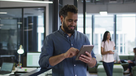 Smiling-mixed-race-businessman-standing-in-office-using-tablet-and-looking-up-to-camera