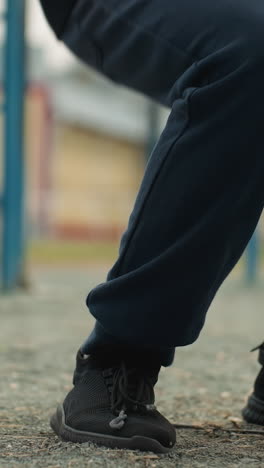 close-up of legs in motion as someone performs a workout, squatting up and down near an iron pole, a blurred yellow building and a person walking in the background