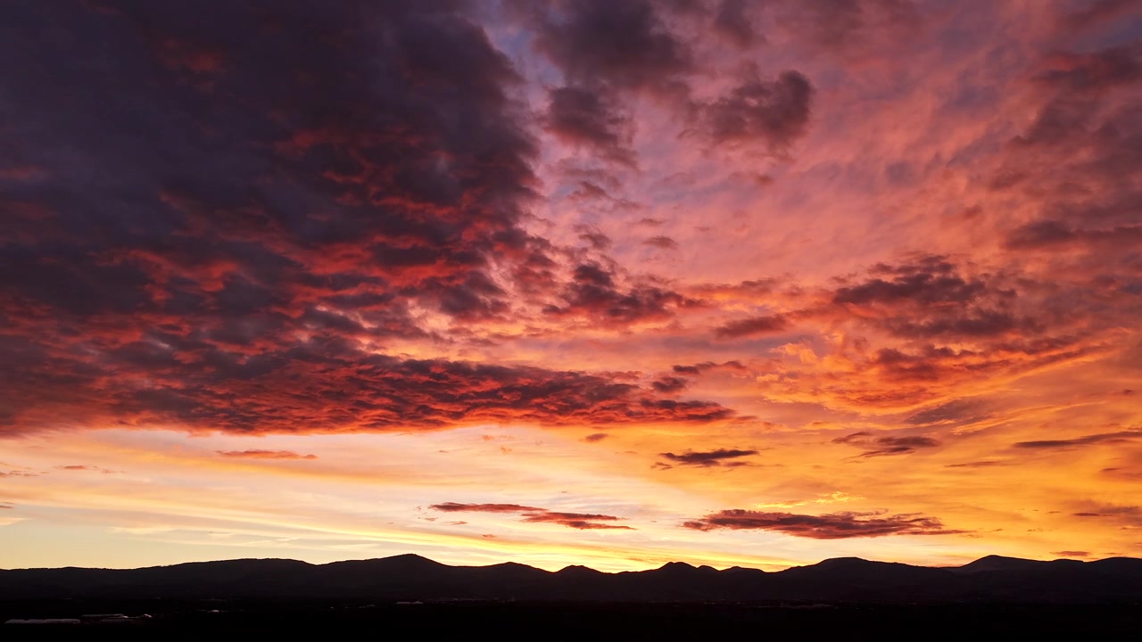 Timelapse aéreo 4K con dron capturando un manto de nubes ardientes en el estallido final del atardecer. Tonos brillantes de naranja y rojo brillan intensamente antes de desvanecerse en el crepúsculo sobre la silueta de una montaña