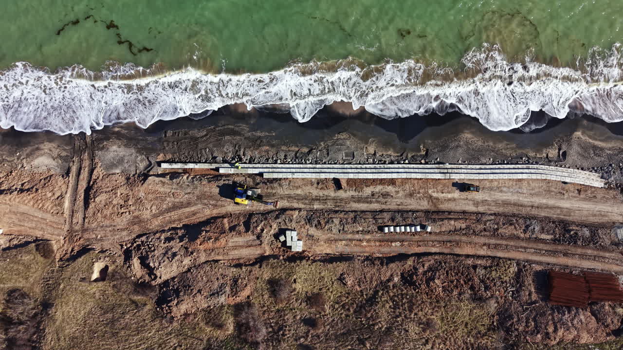 Lavori di costruzione lungo la costa vicino a una spiaggia, con attrezzature e materiali visibili a terra durante le ore diurne.