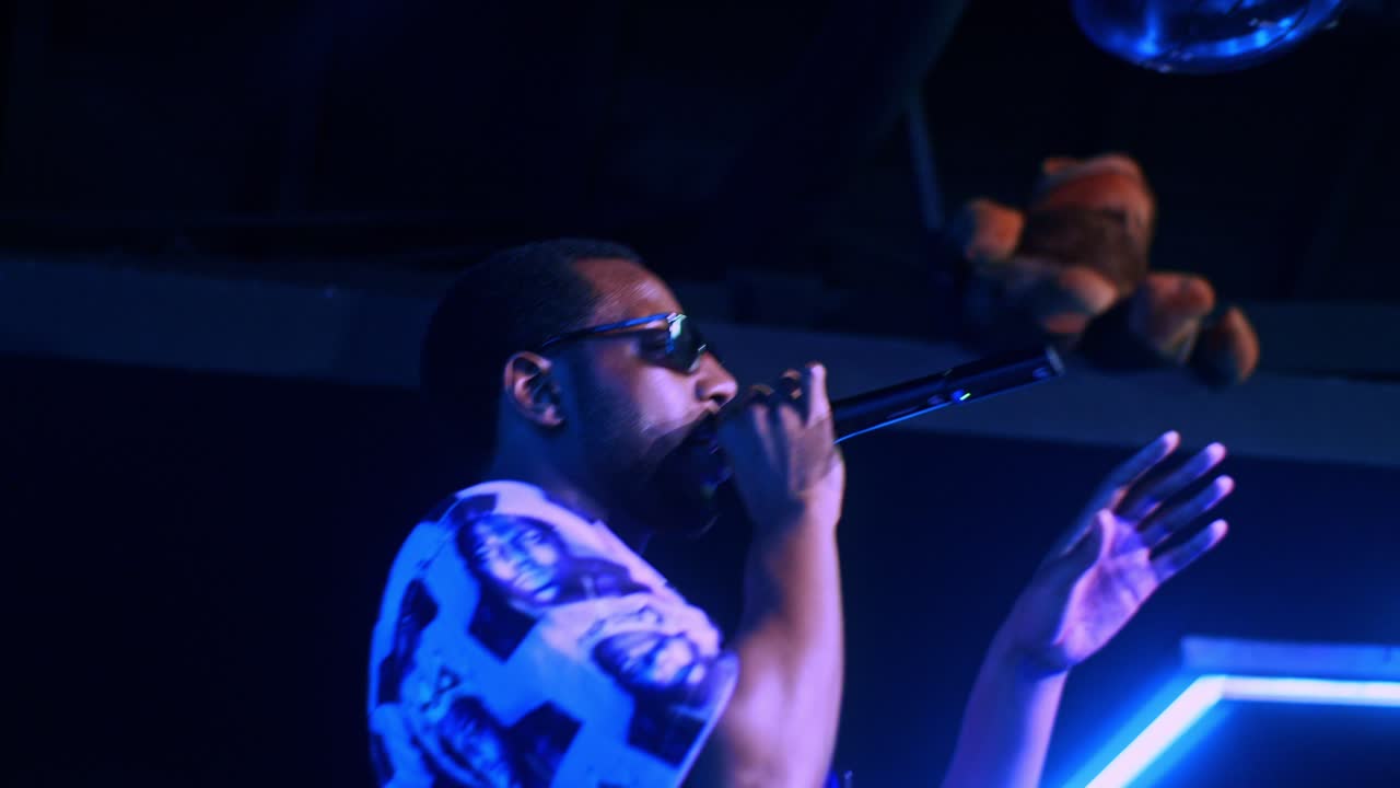Black african male man artist singer rapper showman performs concert party on the club stage in dark scenery with neon lights blue theme and makes speech
