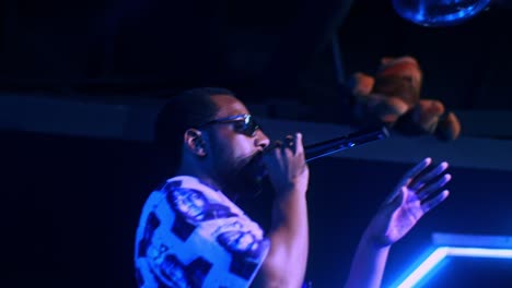 Hombre-Africano-Negro-Artista-Cantante-Rapero-Showman-Realiza-Una-Fiesta-De-Concierto-En-El-Escenario-Del-Club-En-Un-Escenario-Oscuro-Con-Luces-De-Neón-Tema-Azul-Y-Hace-Un-Discurso