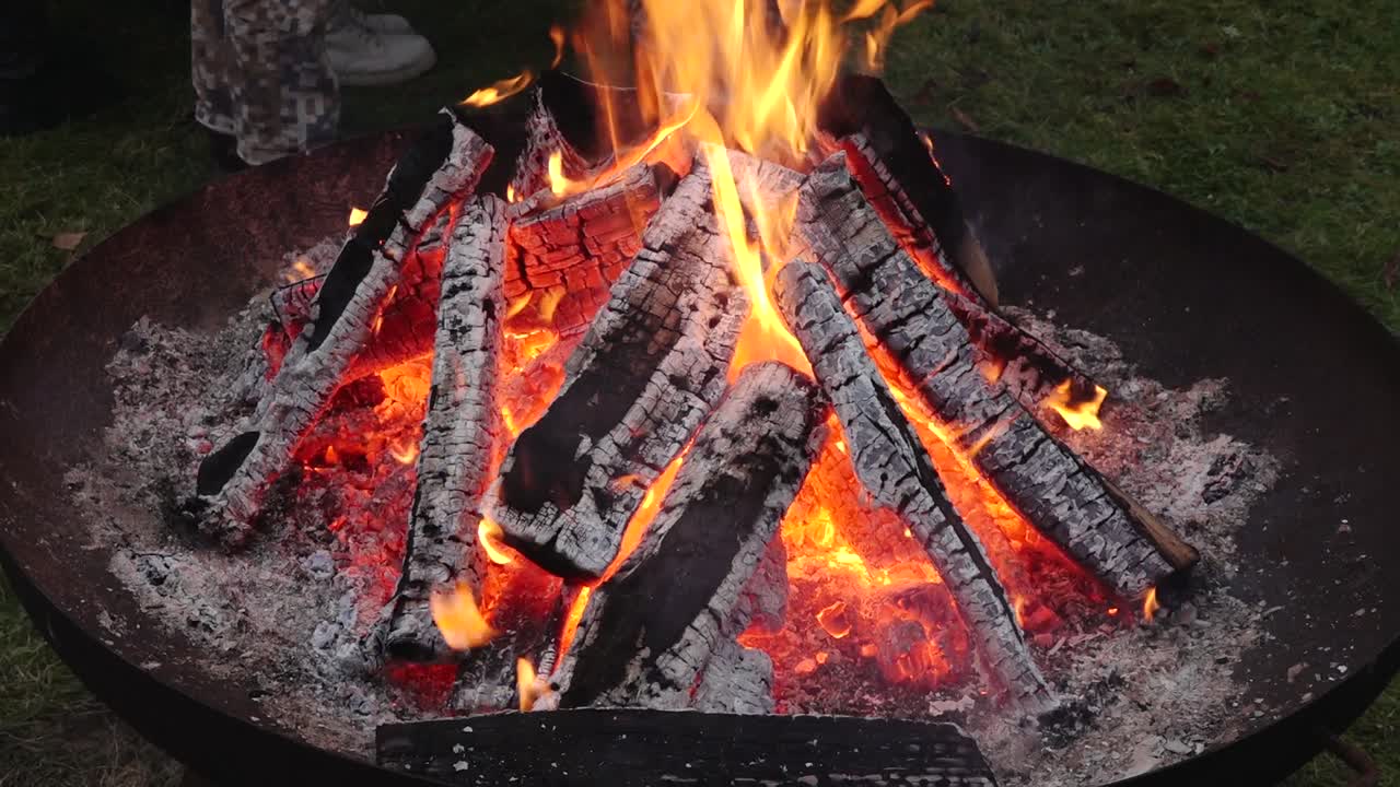A burning bonfire with glowing logs at night, surrounded by people in warm clothing during a solemn gathering. The scene captures a sense of community and reflection.
