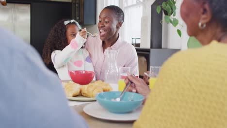 video of happy african american daughter laughing and feeding mother at family breakfast table
