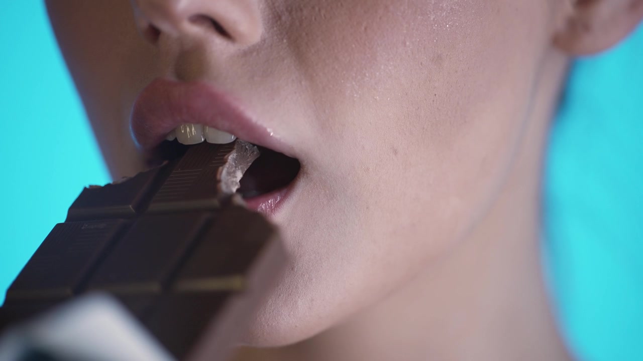 Close-Up Of Woman's Mouth Eating Chocolate Bar Against Blue Background, Studio Shot