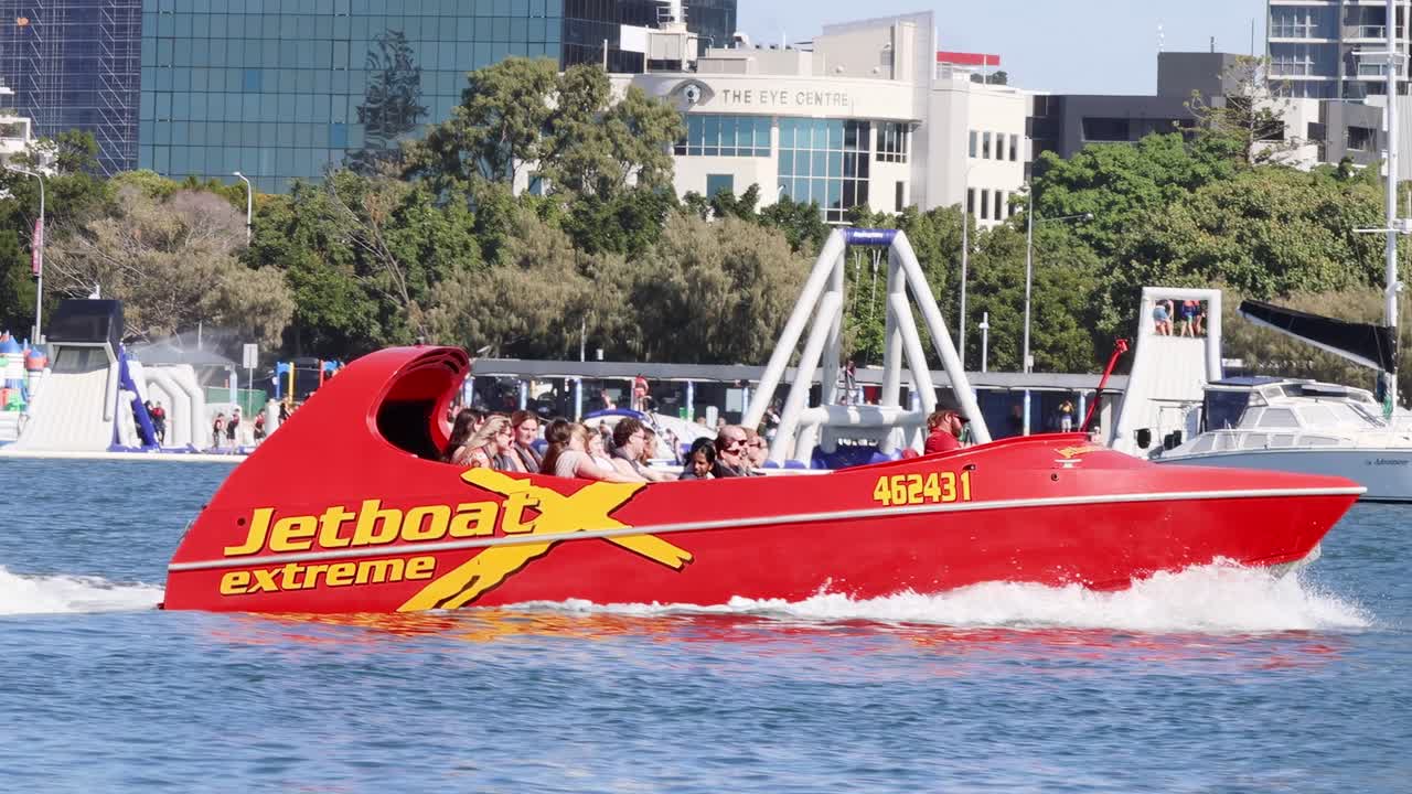 A vibrant red jet boat speeds along the water with city buildings in the background.