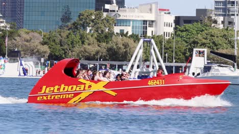 a vibrant red jet boat speeds along the water with city buildings in the background.