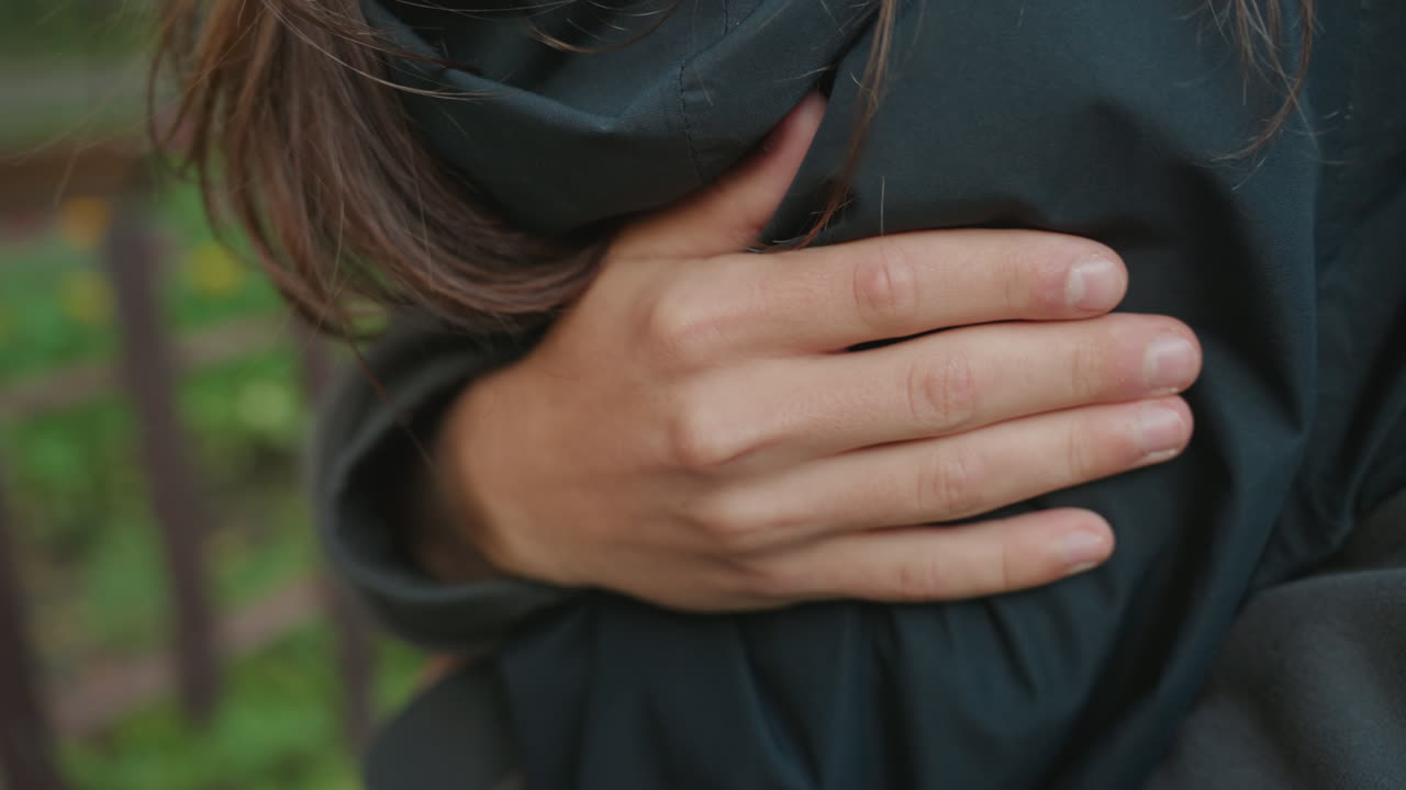 Primo piano di una mano appoggiata sul petto, abbraccio confortante vicino alla recinzione del giardino con palmo e dita visibili che premono delicatamente, dettaglio tattile rilassante e manica di giacca comoda, gesto di supporto intimo che trasmette
