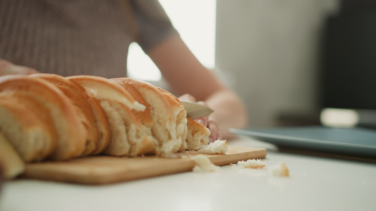Close up of chef slicing loaf of bread with knife on wooden board focus on serrated blade cutting through crust into even slices with crumbs falling on white table under soft window light in kitchen
