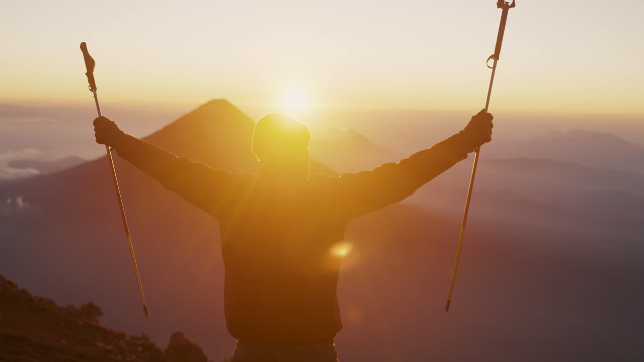 un hombre levantando sus brazos con palos de senderismo sobre el pico de acatenango frente al volcán agua durante la puesta de sol en guatemala, américa central