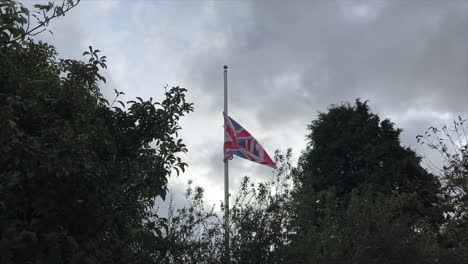 Slow-motion-half-mast-UK-Union-Jack-Flag,-between-trees,-fluttering-in-a-light-breeze