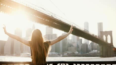 back view of happy tourist girl with flying hair enjoying brooklyn bridge view in new york from embankment, arms open