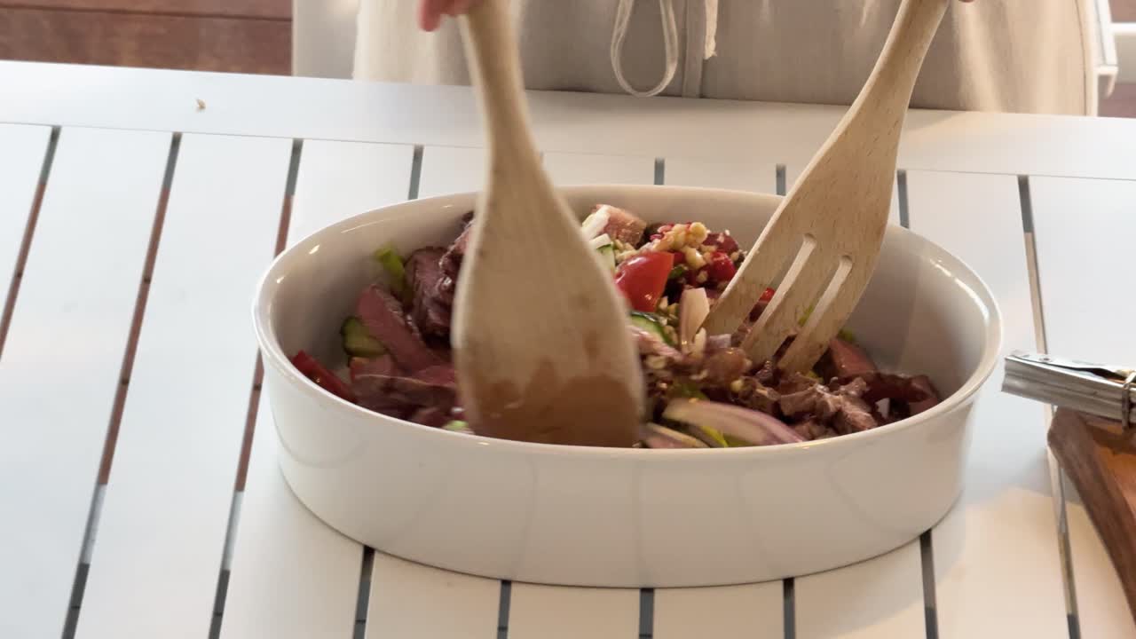 Person uses wooden utensils to toss a colorful Thai beef salad with fresh vegetables in a white bowl on a bright kitchen table