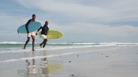 african american father and teenage son running on a beach holding surfboards and talking