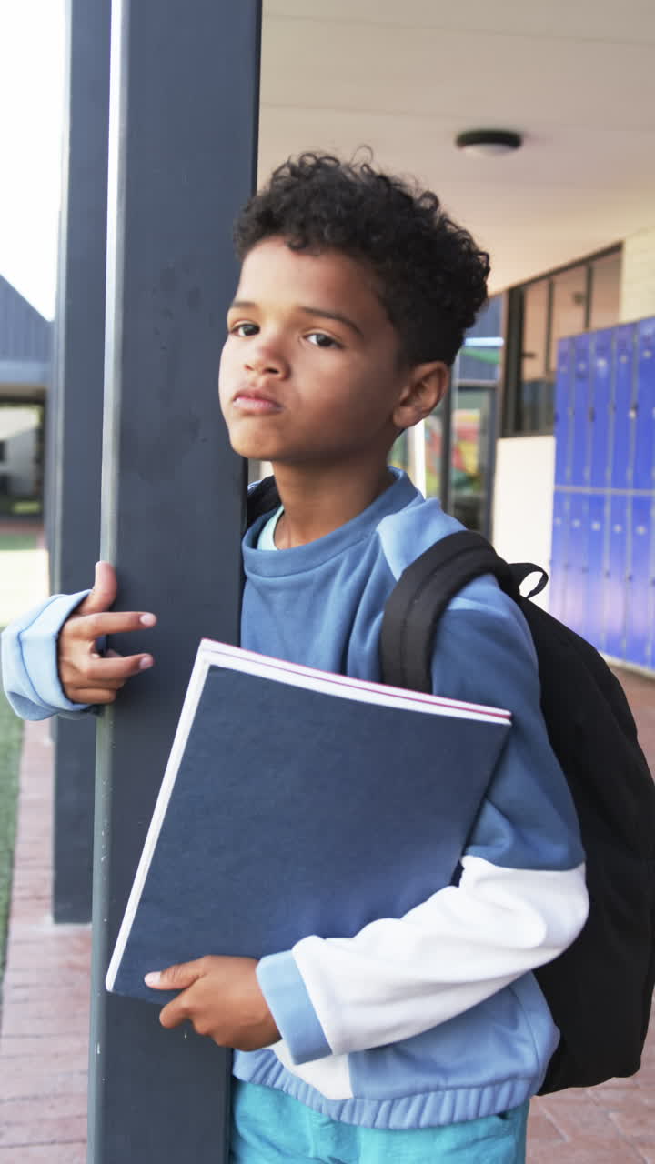 Vertical video: In school, boy holding notebook and leaning against pole, wearing backpack