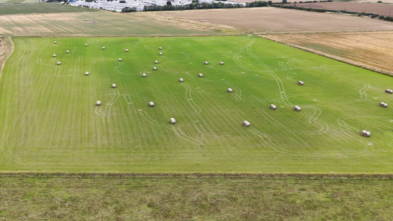 Drone footage glides over a vibrant green field dotted with round hay bales, surrounded by farmland under natural daylight, capturing rural harvest scenery