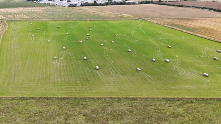 Drone footage glides over a vibrant green field dotted with round hay bales, surrounded by farmland under natural daylight, capturing rural harvest scenery