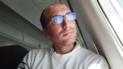 a serious man with glasses looks out of an airplane window