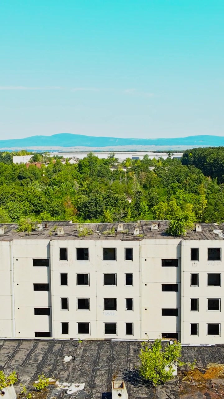 Filmagem aérea em vídeo vertical mostra edifícios de concreto abandonados com janelas quebradas e vegetação no telhado, cercados por floresta exuberante e colinas distantes.