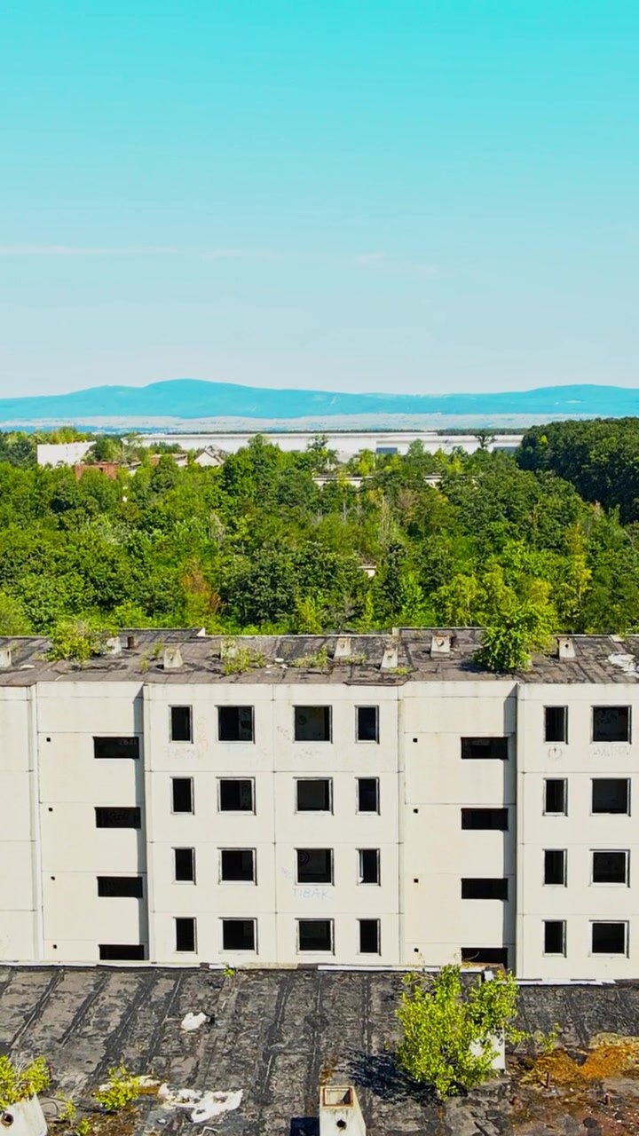 Video verticale con riprese aeree che mostrano edifici in cemento abbandonati con finestre rotte e vegetazione sui tetti, circondati da una foresta lussureggiante e colline distanti.