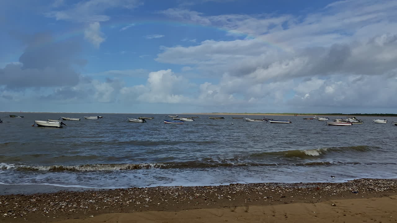 Alguns barcos de pesca flutuam em águas agitadas sob um tênue arco-íris que se estende por um céu costeiro brilhante, mas instável.