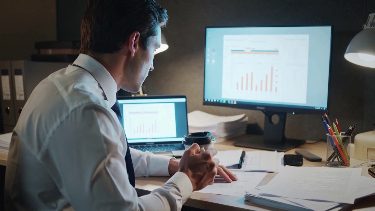 Seeing bar chart on dual screens, analyst in shirt, tie scanning reports, sipping coffee at desk