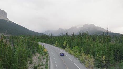 aerial dolly in of car driving on road surrounded by pine trees, canadian rockies in background, banff national park, alberta, canada