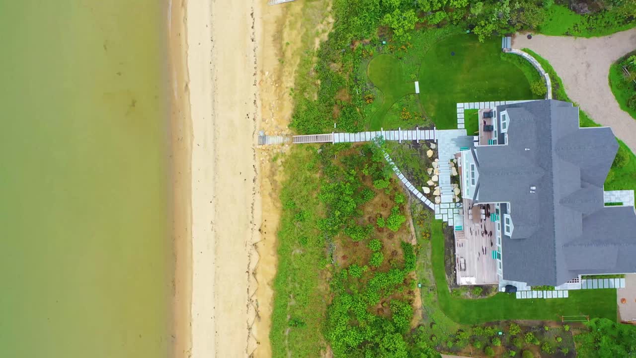 Overhead aerial drone footage of seaside residential community with houses, sandy shoreline, calm ocean waters, and lush trees surrounding beachfront neighborhood streets