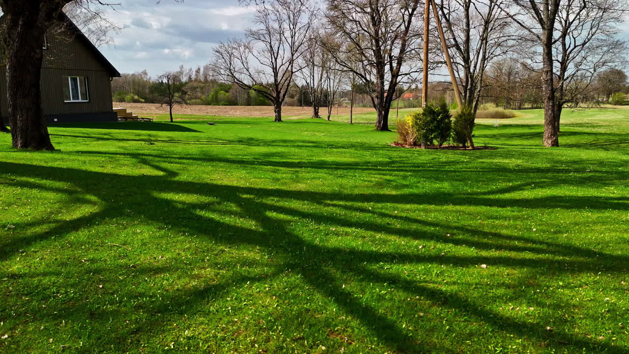 A slow, forward-moving drone shot glides across a vibrant green lawn in the yard of a country house in Latvia during a sunny spring day, casting long shadows from the trees.