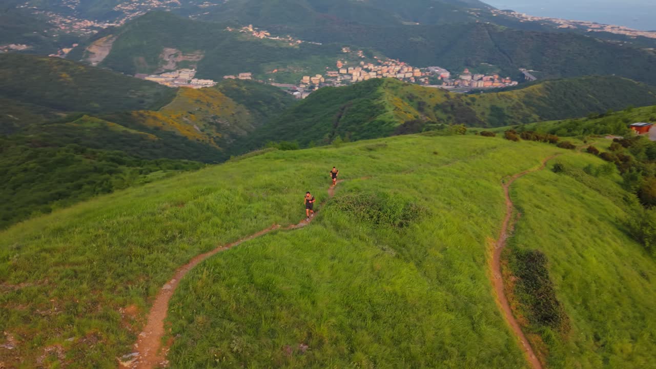 Two men trail running on a lush green mountain path in Italy, slow motion drone shot