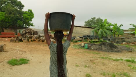 After-fetching-some-water,-a-village-woman-is-carrying-a-heavy-container-with-water-on-top-of-her-head-in-her-neighborhood-in-Kumasi,-Ghana,-in-Africa