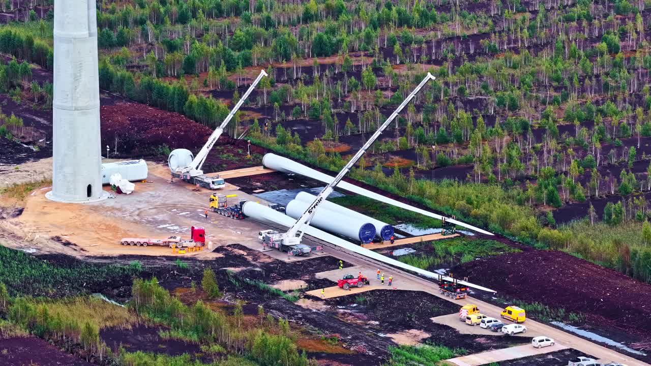 Construction Of A Wind Turbine With Tower Base And Rotor Blades On The Ground Surrounded By Cranes And Machinery. - aerial shot