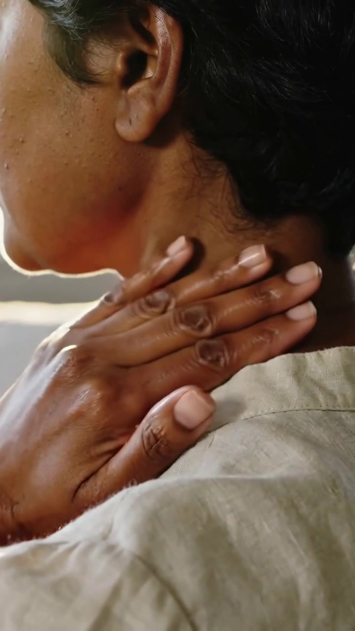 Vertical video: Turning, man in light shirt rubbing neck with right hand at lit window, easing pain