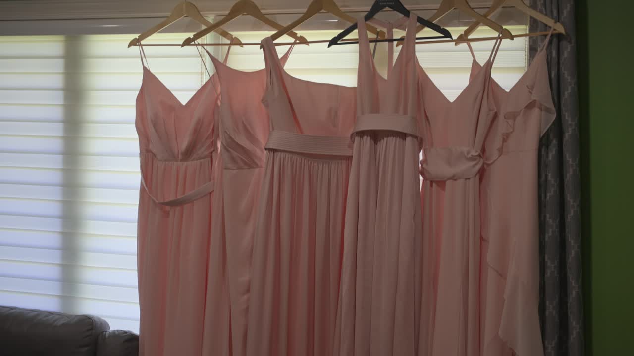 Una fluida ripresa in tilt-down che mostra sei abiti da damigella rosa tenue in vari stili, appesi ordinatamente a un'asta per tende davanti a una finestra mentre il corteo nuziale si prepara per il grande giorno.