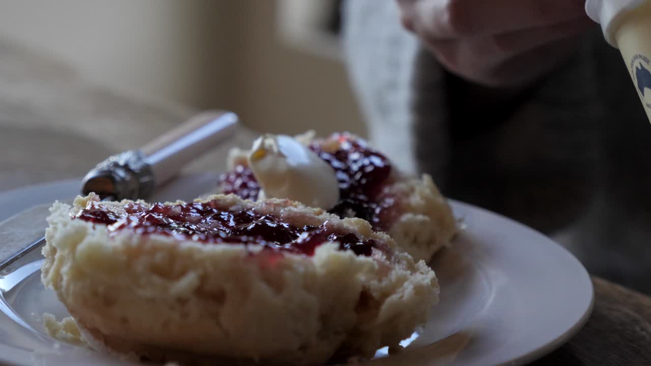 칼로 접시에 잼으로 덮인 갓 구운 수제 전통 스콘에 두꺼운 흰색 크림의 여성 손 숟가락 덩어리