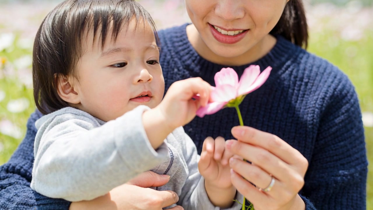 紺色のセーターを着た母親が、牧草地でピンクの花びらに触れる幼児を導き、探索させている