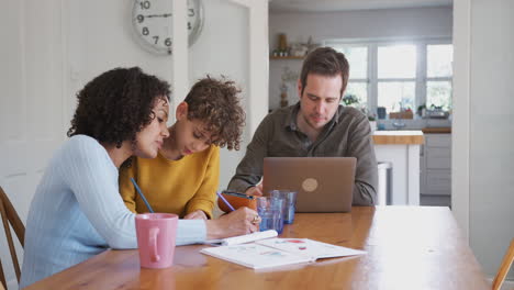 el padre trabaja en la computadora portátil mientras la madre ayuda al hijo con la tarea en la mesa de la cocina