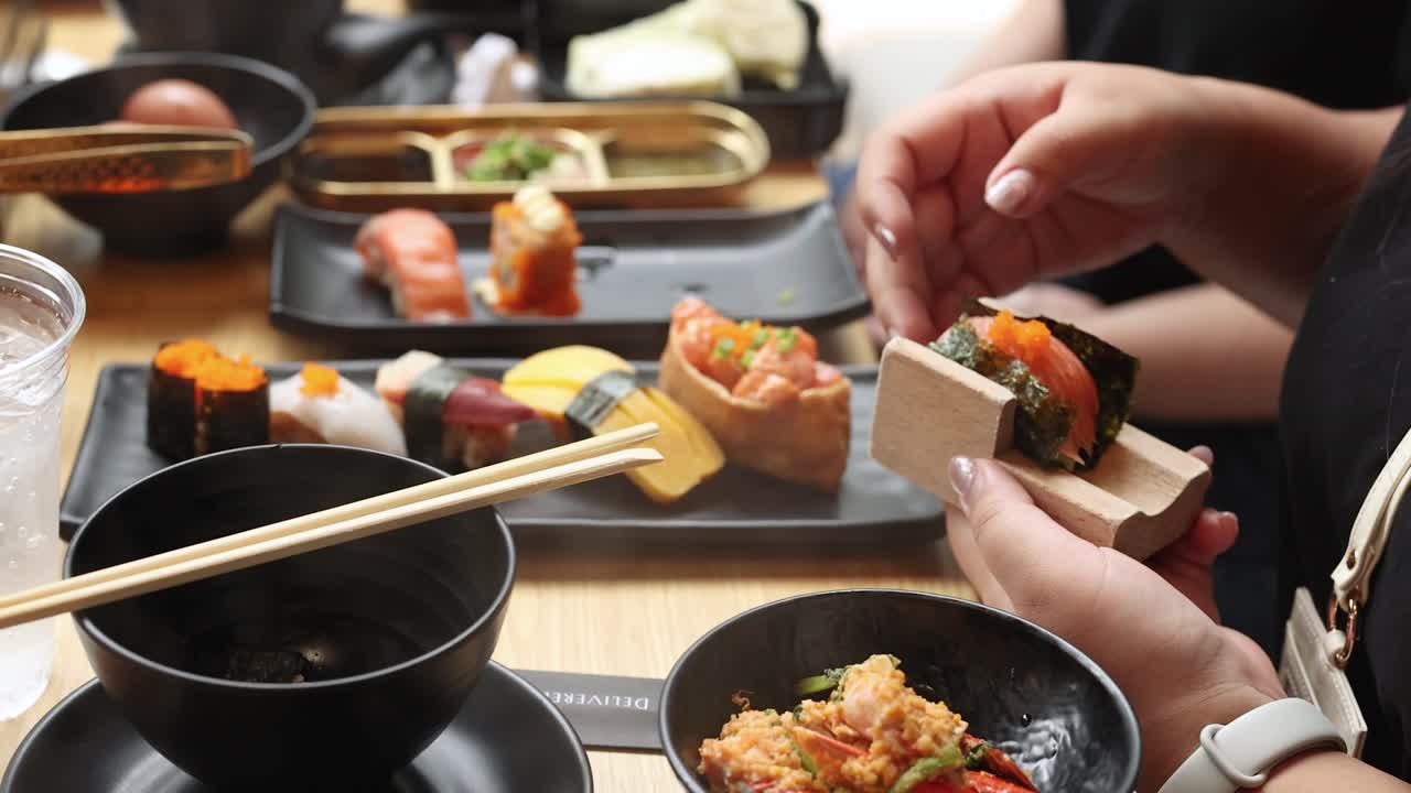 Person picks fresh assorted nigiri sushi with chopsticks in bright, casual Japanese restaurant setting