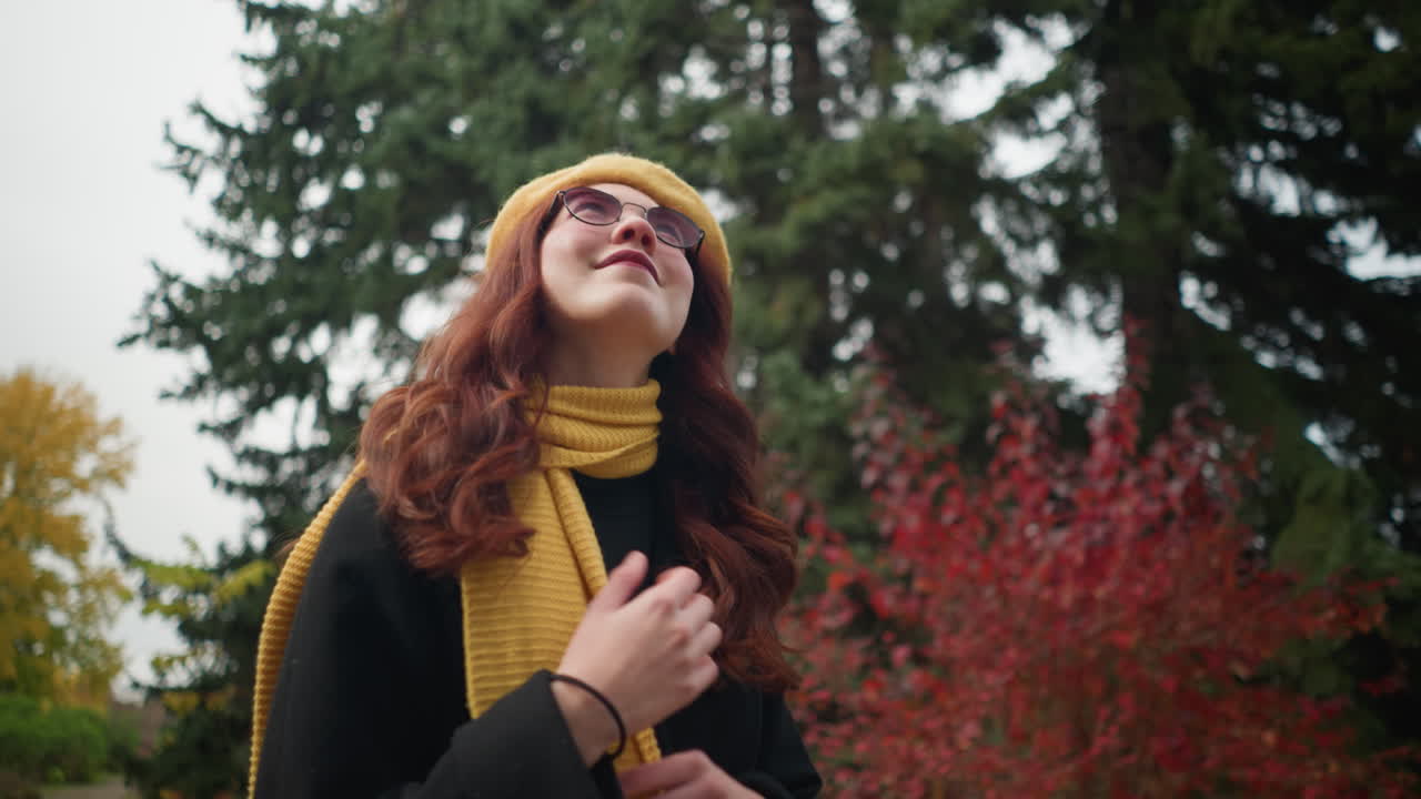 긴 물결 모양의 은 머리카락을 가진 우아한 여인이 검은 코트와 노란색 베레트를 입고 스카프와 일치하는 스카프를 만지며, 다채로운 가을 잎자루로 장식된 활기찬 공원을 고 있습니다.