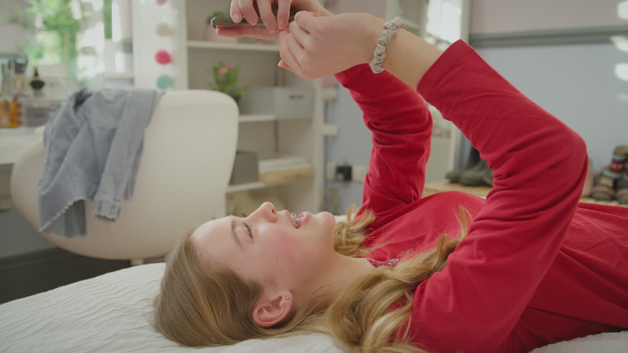 집에서 침대에 누워 얼굴을 당기고 드폰으로 셀피를 찍는 정형 치과 브레이크를 착용한 십대 소녀 - 느린 동작으로 촬영