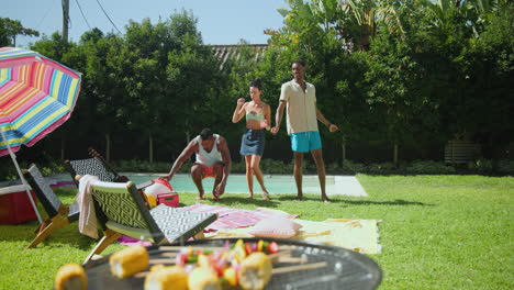 diverse friends dancing joyfully by poolside, enjoying summer day with barbecue nearby