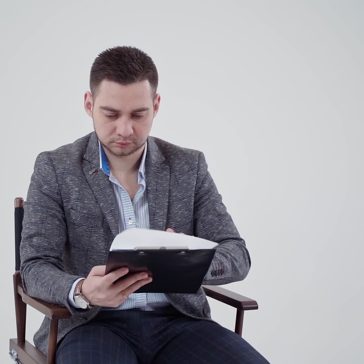 Handsome man in grey costume holding a folder. Young businessman sitting in the chair and reading documents. Slow motion.