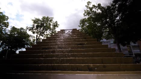 Desde-Lo-Alto-De-Las-Escaleras,-Una-Fuente-Cae-En-Cascada,-Vertiendo-Con-Gracia-A-Los-Lados-De-Los-árboles