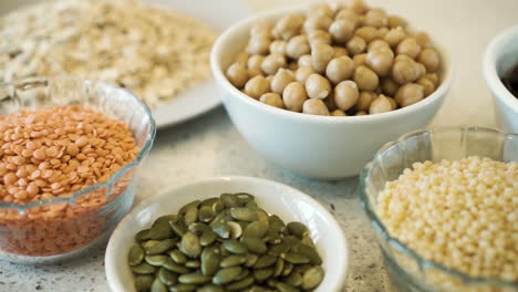 Various-legumes,-seeds-and-grains-in-bowls-on-white-bench,-pan-close-up